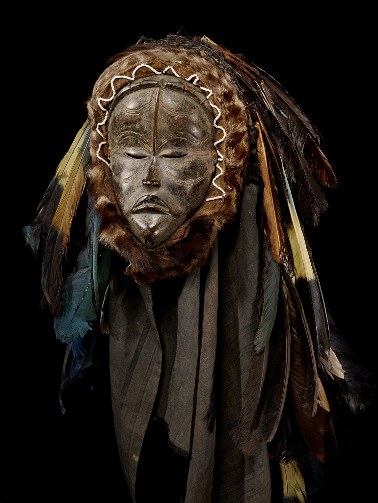 A mask with closed eyes and a slight frown. In place of hair, the mask has many long and multi-colored feathers, and what appears to be a fur trim around the edges of the face.