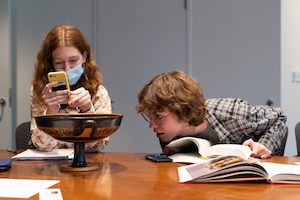 Two people are seated near a large, shallow terracotta cup with handles, a tall stem, and decoration in red against a black background. The person on the right of the vase is leaning forward intently to look at the underside of the cup, holding a book open as he does so. The person on the left is slightly behind the cup, and is holding up a phone as if they might be taking pictures or reading from the screen.