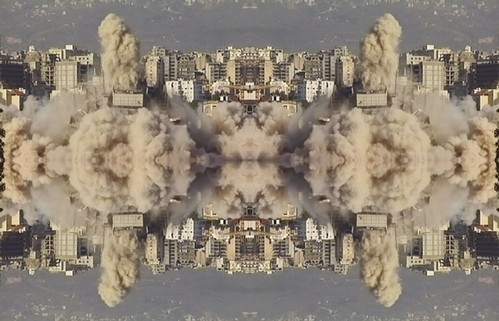 A repeated kaleidoscopic image depicts beige and white residential buildings against a blue and grey mountainous backdrop with beige plumes of smoke and dust emerging from center. The image is mirrored over the center horizon.