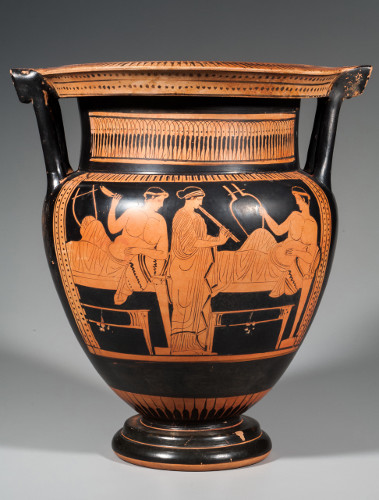 A vessel with two column-like handles has ornamental patterns around the neck and on the wide, flattened rim. The scene on the visible side of the bowl's body depicts two young men reclining on couches and a stand woman playing an oboe-like instrument.