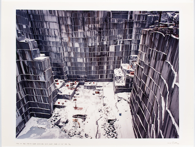 A photograph with muted gray colors shows a stone quarry from high up, looking down at the floor and three sides of a roughly rectangular space. Stone has been removed from the quarry walls in horizontal rows, and combined with the muted gray of the rock it gives the impression of high-rise apartments. Far below at the floor of the quarry, the bright orange of heavy machinery offers spots of color.