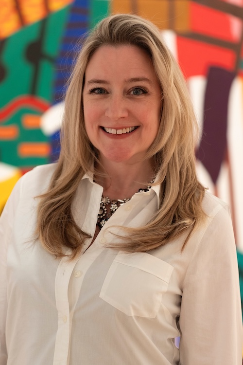 Mariah Keller, a smiling blond woman, looks out at the camera. She is standing in front of a colorful abstract artwork.