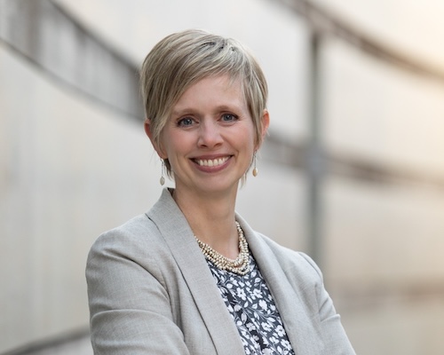 A portrait of a light-skinned woman with fair hair. She is smiling widely and looking directly at the camera.
