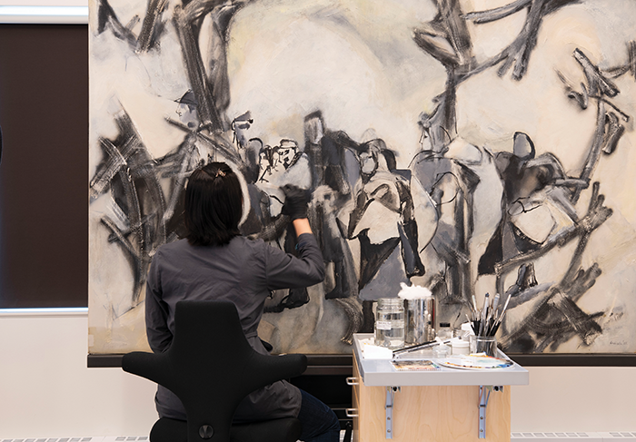 A dark-haired person with their back to the camera works on cleaning a large abstract painting.