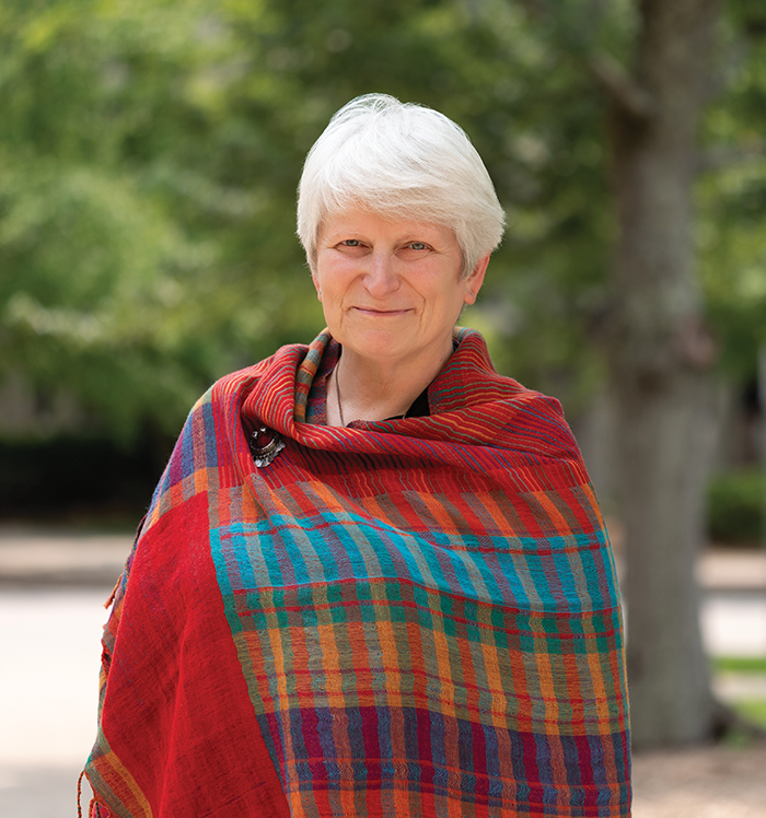 A white haired person looks out at the camera. They have a colorful shawl wrapped around their shoulders and torso.