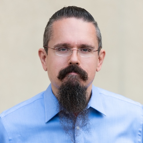 A black and white headshot of a man with glasses, slicked-back dark hair, and a dark mustache and beard.