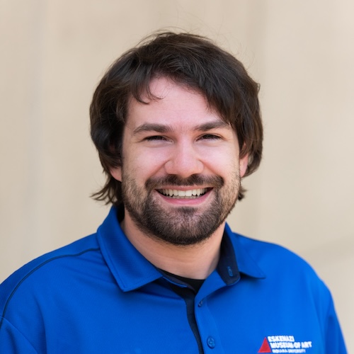 A black and white headshot of a man with dark hair smiling out at the viewer. He has a short beard and a short shag haircut.