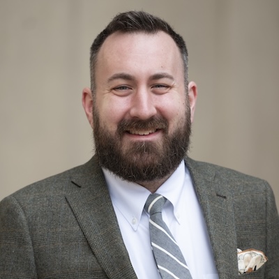 A smiling man with a beard and short hair. He wears a grey suit and tie with a patterned pocket square.