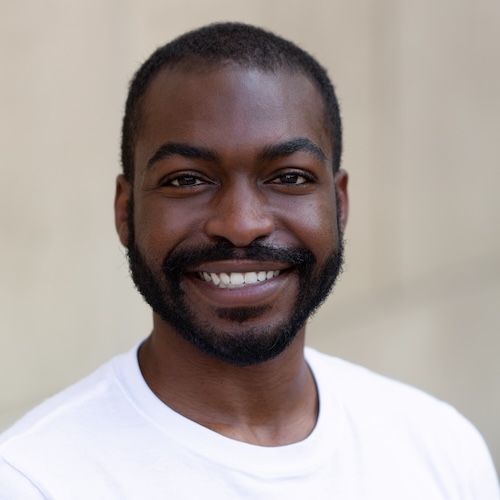 A black and white headshot of a young man with short dark hair and a short beard smiling widely.