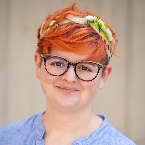 A black and white headshot of a person with rounded glasses and very short hair tied with a scarf, who is looking directly at the camera and smiling slightly.