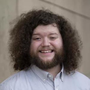 A young man with long, curly, light brown hair and a short beard smiles at the camera.
